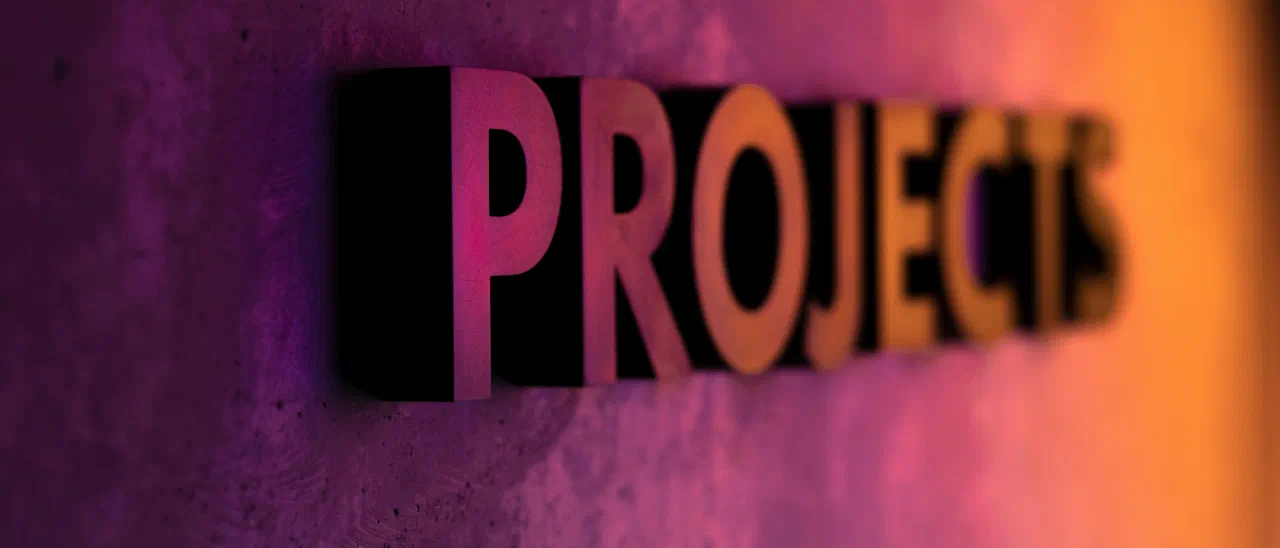 Plastische Buchstaben an einer Betonwand bilden das Wort PROJECTS, angestrahlt in Pink und Orange