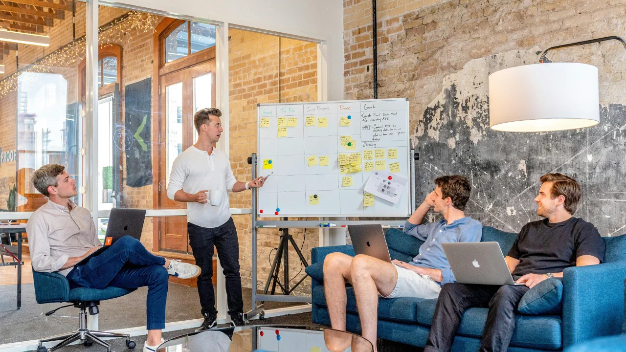 Ein Mann erklärt vor einem Whiteboard mit Aufgabenkarten, vier Zuhörer mit Laptops sitzen im Halbkreis davor