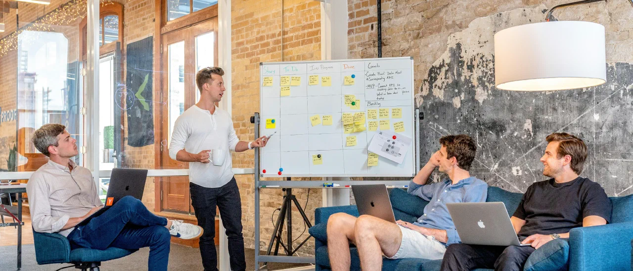 Ein Mann erklärt vor einem Whiteboard mit Aufgabenkarten, vier Zuhörer mit Laptops sitzen im Halbkreis davor